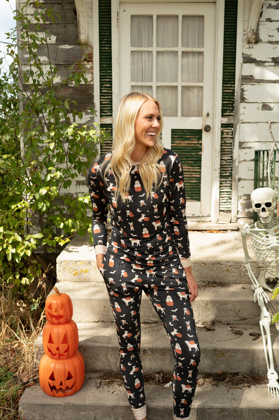 Woman wearing black Pumpkin Pups fabric pajamas with dog and ghost prints, standing by a skeleton and plastic pumpkins.