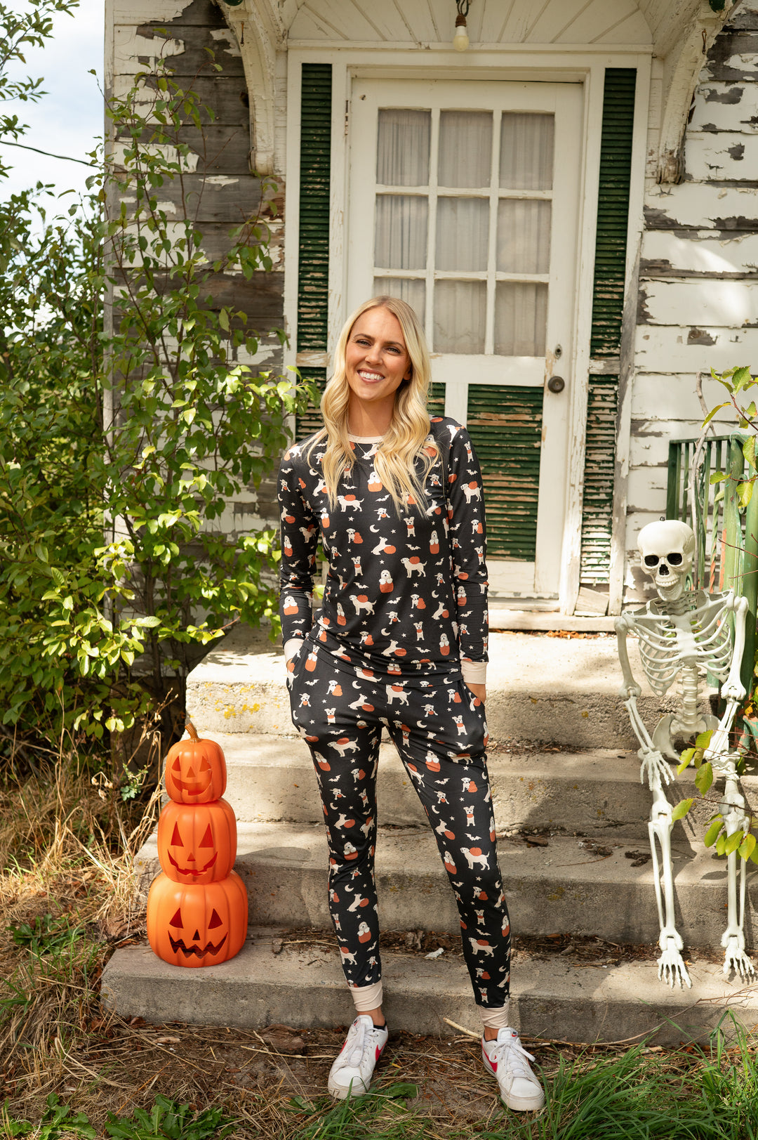 Woman in black Pumpkin Pups pajamas with dog and pumpkin prints, standing by a skeleton and jack-o&