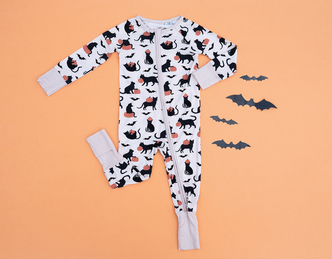 White fabric zip footie with black cat and orange pumpkin print, laid flat on orange background with four paper bats.