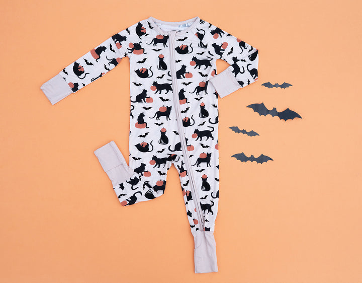 White fabric zip footie with black cat and orange pumpkin print, laid flat on orange background with four paper bats.