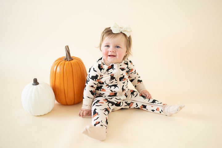 Baby in Pumpkin Cats zip footies sitting next to one orange and one white pumpkin.